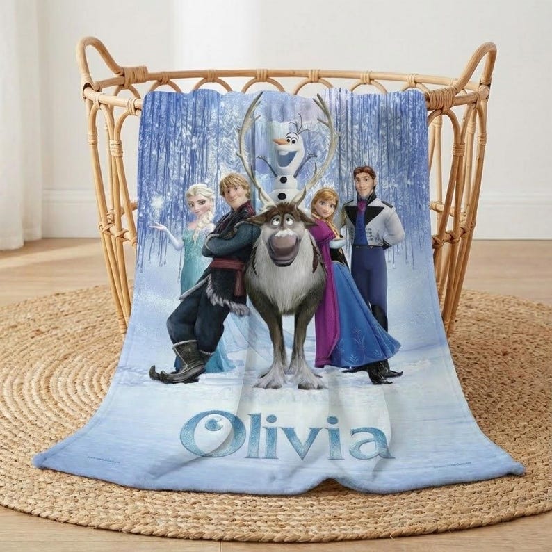 Puede incluir: Una manta azul con personajes de la pel&iacute;cula animada Frozen, incluyendo a Elsa, Anna, Kristoff, Sven, Olaf y Hans. La manta se muestra en una cesta de mimbre y tiene el nombre "Olivia" impreso.