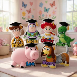 May include: A collection of Toy Story character cutouts, each wearing a graduation cap and personalized with a name. Characters include Woody, Buzz Lightyear, Jessie, Rex, Hamm, and Slinky Dog. The cutouts are in a child's bedroom.