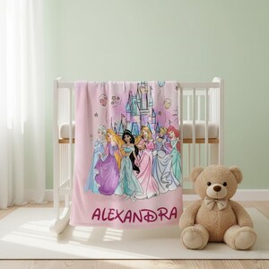 May include: Pink fleece blanket featuring a group of cartoon princesses and a castle illustration. The name "ALEXANDRA" is printed in a matching pink font. The blanket is draped over a white crib, with a teddy bear sitting nearby.
