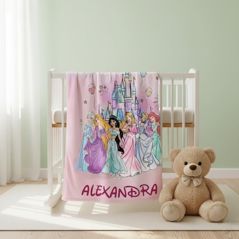 May include: Pink fleece blanket featuring a group of cartoon princesses and a castle illustration. The name "ALEXANDRA" is printed in a matching pink font. The blanket is draped over a white crib, with a teddy bear sitting nearby.