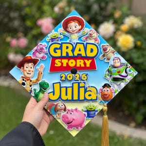 May include: A graduation cap featuring characters from the Toy Story movie. The cap is decorated with the words "GRAD STORY 2026 Julia" in yellow and blue. The cap has a gold tassel.