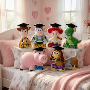 May include: A collection of plush Toy Story character figures, each wearing a graduation cap and personalized with a name. The figures include Woody, Buzz Lightyear, Jessie, Rex, Hamm, and Slinky Dog, set on a bed with pink accents.