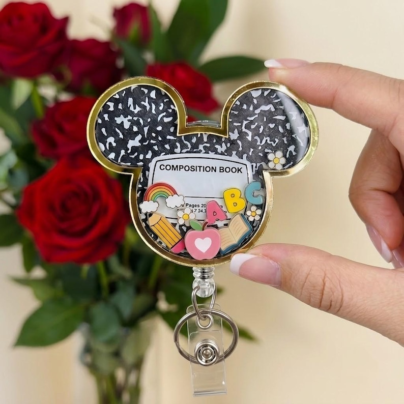 May include: A Mickey Mouse-shaped badge reel featuring a black and white composition book pattern. The center includes a white label with the text COMPOSITION BOOK and colorful miniature charms of a rainbow, pencil, apple, book, and letters A, B, and C.