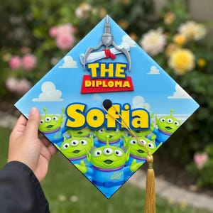 Op de afbeelding: Een afstudeerhoed met een blauwe lucht, witte wolken en de tekst "THE DIPLOMA". Het heeft een grijpmachine-afbeelding en de naam "Sofia" in geel. Groene alien-personages zijn ook aanwezig.