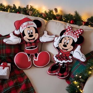 Puede incluir: Dos cojines de peluche con forma de Mickey Mouse y Minnie Mouse vestidos con ropa festiva. Mickey lleva una camisa de cuadros rojos y negros y un gorro de Papá Noel con el nombre Sophia. Minnie lleva un vestido y un lazo de cuadros con el nombre Olivia.