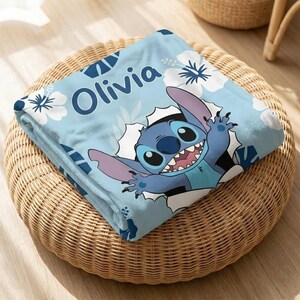 May include: A light blue blanket featuring the character Stitch bursting through a white hole. The blanket has white floral and blue geometric patterns, with the name "Olivia" in blue. The blanket is folded on a woven wicker ottoman.