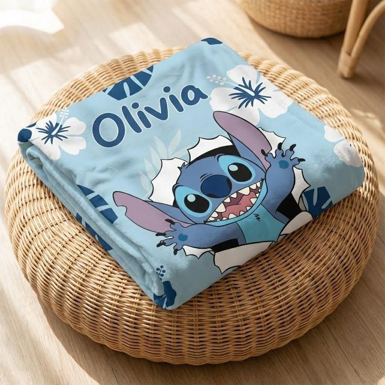 May include: A light blue blanket featuring the character Stitch bursting through a white hole. The blanket has white floral and blue geometric patterns, with the name "Olivia" in blue. The blanket is folded on a woven wicker ottoman.