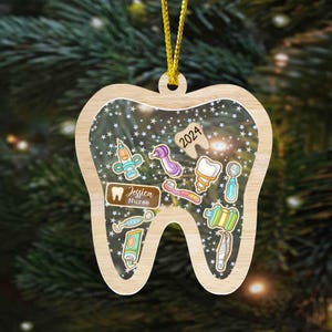 May include: Wooden Christmas ornament shaped like a tooth, featuring dental tools and the text "2024." The ornament includes the name "Jessica Nurse" on a small plaque. A gold string is attached for hanging.