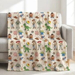 May include: A beige throw blanket featuring a repeating pattern of characters from the animated film Toy Story. The design includes Woody, Buzz Lightyear, Rex, Mr. Potato Head, and other iconic elements in a colorful arrangement.