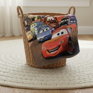 Puede incluir: Una cesta de mimbre tejida con asas de cuerda contiene una colorida manta con personajes de la pel&iacute;cula de animaci&oacute;n "Cars". La manta muestra a Rayo McQueen y otros personajes de coches. La cesta est&aacute; sobre una alfombra redonda blanca.