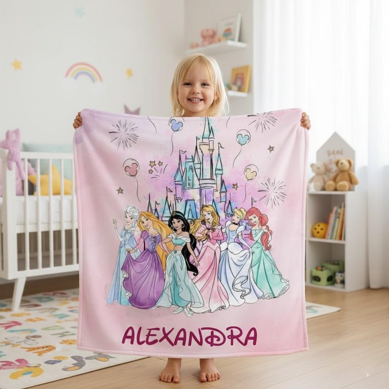 May include: A pink blanket featuring a castle and several cartoon princesses. The blanket has the name "ALEXANDRA" printed in a dark pink script. The princesses are wearing colorful gowns. The background has fireworks and balloons.