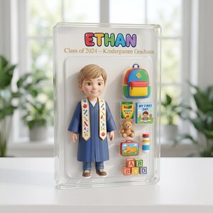 May include: A commemorative display featuring a kindergarten graduate figurine in a blue gown. The display includes a backpack, books, a teddy bear, a lunchbox, and alphabet blocks. The text "ETHAN Class of 2024 - Kindergarten Graduate" is displayed.