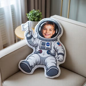 Könnte beinhalten: Kissen in Astronautenform mit dem lächelnden Gesicht eines Kindes im Helm. Das Kissen ist weiß und grau, mit NASA- und amerikanischer Flaggen-Details. Der Astronaut hält eine weiße Flagge.