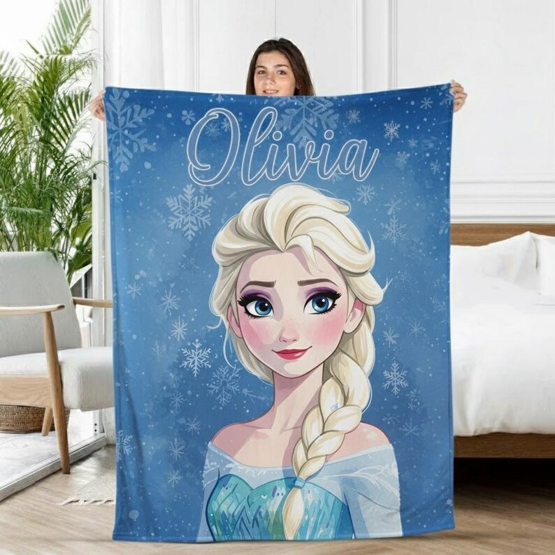 Pode incluir: Um cobertor azul com uma ilustra&ccedil;&atilde;o de desenho animado de uma mulher loira com olhos azuis e uma tran&ccedil;a, rodeada por flocos de neve. O nome "Olivia" est&aacute; escrito em it&aacute;lico no topo.