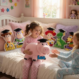 May include: Two young girls in pajamas are sitting on a bed, smiling and holding a large pink piggy bank with the name "Leo" on it. Behind them are character-shaped pillows of Woody, Buzz Lightyear, Jessie, Rex, and Slinky Dog, each wearing a graduation cap.