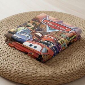 Puede incluir: Manta plegada con personajes de la pel&iacute;cula de animaci&oacute;n Cars. La manta muestra a Rayo McQueen y otros personajes en un dise&ntilde;o colorido. El nombre "Daniel" se muestra en un letrero rojo y blanco.