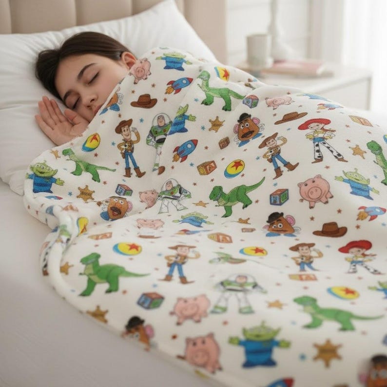 May include: A white blanket featuring colorful cartoon characters, including a green dinosaur, a cowboy, and a space ranger. The blanket is spread over a bed, with a person sleeping underneath.