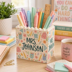 May include: A personalized pen holder with the name "MRS. JOHNSON" on it, filled with colorful pens, pencils, and a wooden ruler. The holder is decorated with school-themed illustrations and floral patterns. A mug with the text "coffee teach repeat" is also visible.