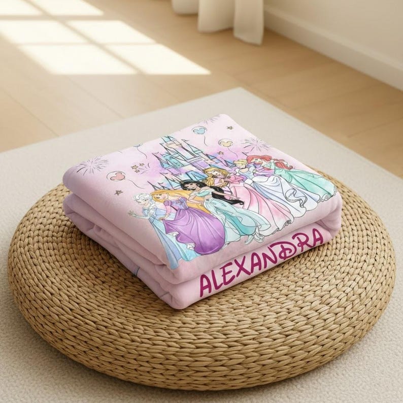 May include: A folded pink blanket featuring a princess castle and several princess characters. The blanket has the name "ALEXANDRA" printed in a cursive font. The blanket is resting on a woven, circular ottoman.