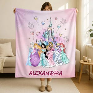 May include: A pink blanket featuring a castle illustration with several princesses in colorful gowns. The blanket has a watercolor effect with balloons and fireworks. The name "ALEXANDRA" is printed in a cursive font at the bottom.