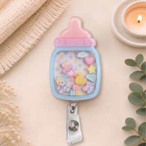 May include: A baby bottle-shaped badge reel featuring a pink cap and light blue base. The clear center contains floating charms including a teddy bear, yellow duck, pacifier, heart, star, clouds, baby socks, and ABC blocks on a polka dot background.