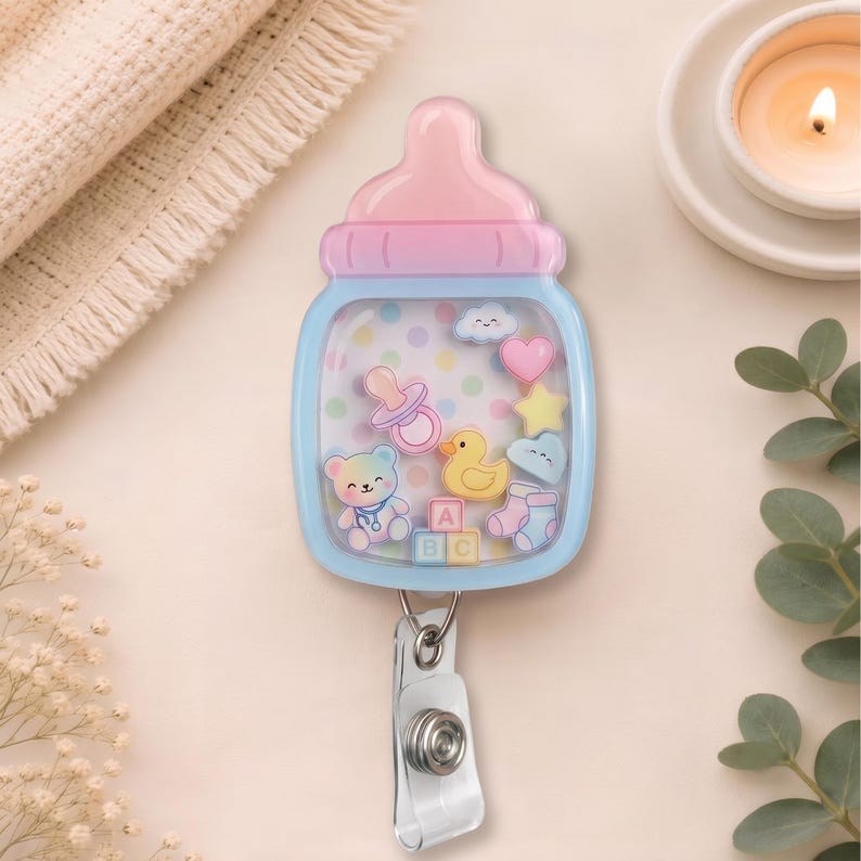 May include: A baby bottle-shaped badge reel featuring a pink cap and light blue base. The clear center contains floating charms including a teddy bear, yellow duck, pacifier, heart, star, clouds, baby socks, and ABC blocks on a polka dot background.