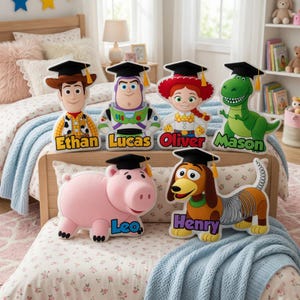 May include: A collection of Toy Story character-shaped pillows, each wearing a graduation cap and personalized with a name. The characters include Woody, Buzz Lightyear, Jessie, Rex, Hamm, and Slinky Dog. The pillows are displayed on a bed with floral bedding.