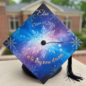 Może przedstawiać: Czapka absolwenta z niebieskim, fioletowym i białym wzorem galaktyki. Czapka zawiera imię "Elsa" oraz tekst "Class of 2026" i "On to my new dream". Czarny chwost wisi z czapki.