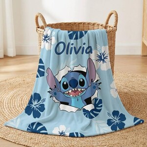 May include: A light blue blanket featuring the character Stitch bursting through a hole, surrounded by white and dark blue floral patterns. The name "Olivia" is printed in blue. The blanket is folded in a wicker basket.