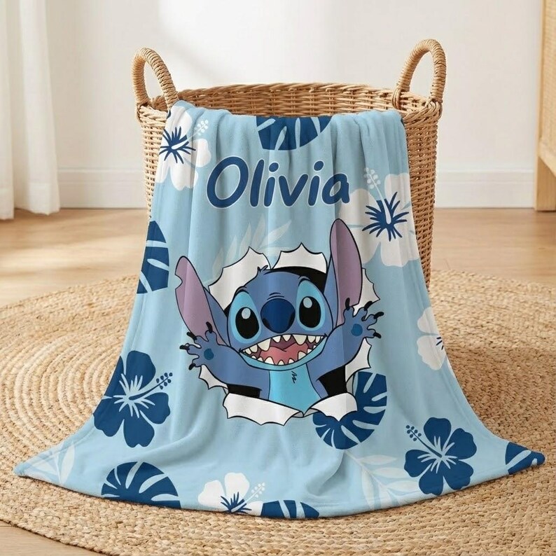 May include: A light blue blanket featuring the character Stitch bursting through a hole, surrounded by white and dark blue floral patterns. The name "Olivia" is printed in blue. The blanket is folded in a wicker basket.
