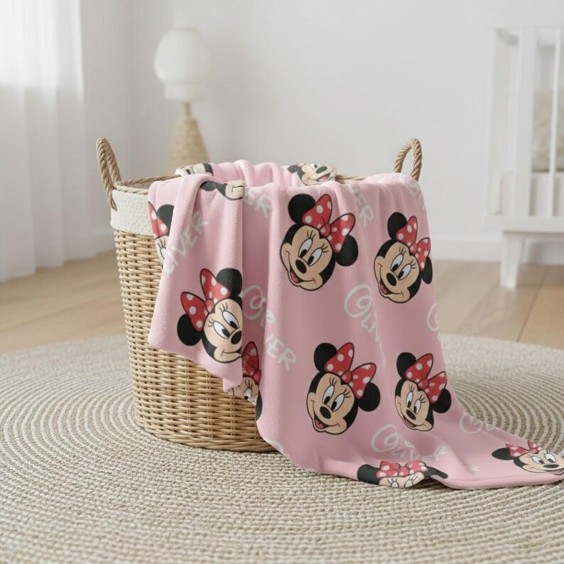 May include: A pink blanket featuring a repeating pattern of Minnie Mouse's face with a red bow and the word "Oliver". The blanket is draped over a wicker basket on a neutral rug.
