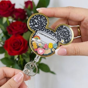 May include: A Mickey Mouse-shaped badge reel featuring a black and white speckled composition book pattern. The center includes a label reading COMPOSITION BOOK with miniature charms of a rainbow, pencil, apple, open book, and the letters ABC.