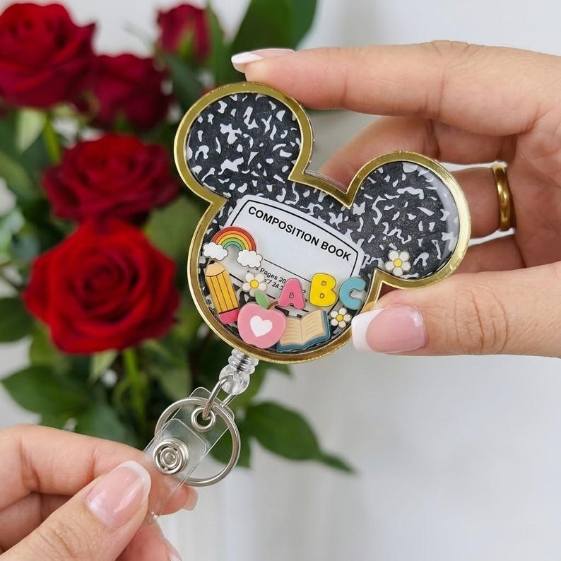 May include: A Mickey Mouse-shaped badge reel featuring a black and white speckled composition book pattern. The center includes a label reading COMPOSITION BOOK with miniature charms of a rainbow, pencil, apple, open book, and the letters ABC.