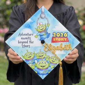 Puede incluir: Un gorro de graduación decorado con el tema de Toy Story. El gorro tiene un fondo de acuarela azul y blanco con el texto "La aventura espera más allá de las estrellas". Incluye una borla dorada y el nombre "Elizabeth".