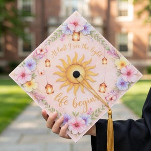Könnte beinhalten: Eine Abschlusskappe mit floralem Design und dem Text "At last I see the light, life begins." Die Kappe zeigt eine Sonnengrafik, hängende Laternen und bunte Blumen in Rosa, Lila und Gelb. Eine goldene Quaste hängt von der Kappe.