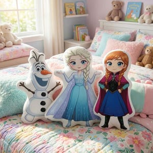 Op de afbeelding: Drie kussens in de vorm van personages uit de Disney-film Frozen: Olaf de sneeuwman, Elsa in een blauwe jurk en Anna in een blauwe jurk en cape. De kussens liggen op een bloemenquilt.