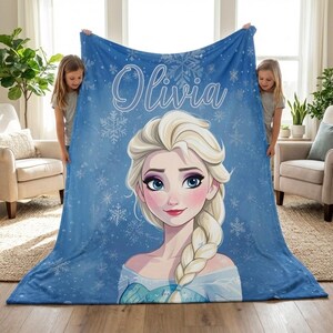 Pode incluir: Um cobertor de l&atilde; azul com uma ilustra&ccedil;&atilde;o de desenho animado de uma princesa com cabelo loiro e olhos azuis. O cobertor tem o nome "Olivia" em escrita branca e desenhos de flocos de neve. O cobertor &eacute; segurado por duas pessoas.