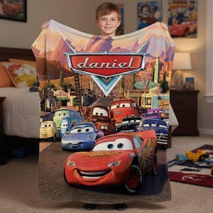 Puede incluir: Una manta de felpa con personajes de la pel&iacute;cula animada Cars. La manta muestra a Rayo McQueen y otros personajes en un paisaje des&eacute;rtico. El nombre "Daniel" se muestra en un emblema rojo y plateado.