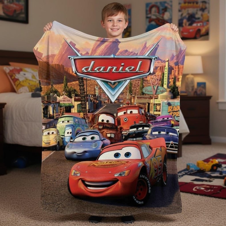 Puede incluir: Una manta de felpa con personajes de la pel&iacute;cula animada Cars. La manta muestra a Rayo McQueen y otros personajes en un paisaje des&eacute;rtico. El nombre "Daniel" se muestra en un emblema rojo y plateado.