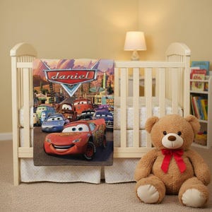 Puede incluir: Una cuna con una manta con tem&aacute;tica de Cars con Rayo McQueen y otros personajes. La manta tiene el nombre "Daniel". Un oso de peluche marr&oacute;n con un lazo rojo est&aacute; sentado junto a la cuna.