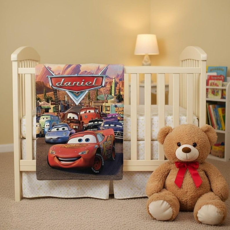 Puede incluir: Una cuna con una manta con tem&aacute;tica de Cars con Rayo McQueen y otros personajes. La manta tiene el nombre "Daniel". Un oso de peluche marr&oacute;n con un lazo rojo est&aacute; sentado junto a la cuna.