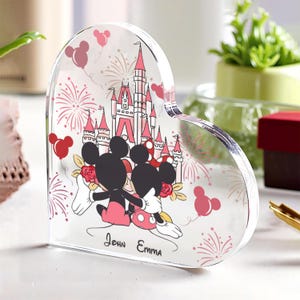 May include: Heart-shaped clear acrylic keepsake with a Disney theme. It features Mickey and Minnie Mouse, a pink castle, fireworks, and balloons. The names "John Emma" are also included. A decorative item.