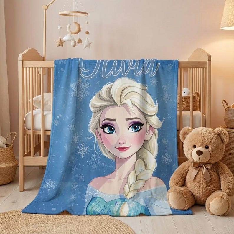 Pode incluir: Um cobertor de l&atilde; azul com uma ilustra&ccedil;&atilde;o de desenho animado de uma princesa, cabelo loiro e olhos azuis. O cobertor tem o nome "Olivia" em escrita branca e detalhes de floco de neve. Est&aacute; sobre um ber&ccedil;o.