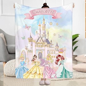 K&ouml;nnte beinhalten: Eine personalisierte Decke mit einem Schloss, Schmetterlingen und vier Prinzessinnen in Kleidern. Die Decke hat ein rosa Banner mit dem Namen "Charlotte". Der Hintergrund ist ein pastellfarbenes Aquarelldesign.