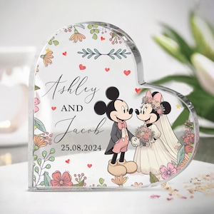 Puede incluir: Un recuerdo de acrílico transparente en forma de corazón con un diseño floral y una imagen de dibujos animados de Mickey y Minnie Mouse vestidos de novia y novio. El texto "Ashley y Jacob 25.08.2024" está impreso en el recuerdo.