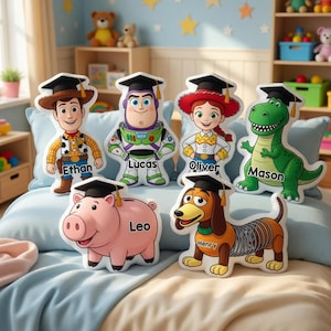 May include: A collection of character-shaped pillows featuring designs from a popular animated movie. The pillows include Woody, Buzz Lightyear, Jessie, Rex, a pig, and a dachshund, each wearing a graduation cap. Names are printed on each pillow.