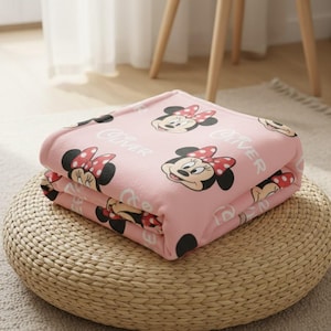 May include: A folded pink blanket featuring a repeating pattern of Minnie Mouse's face with a red and white polka dot bow. The word "Oliver" is printed on the blanket. The blanket is folded and placed on a woven, round ottoman.