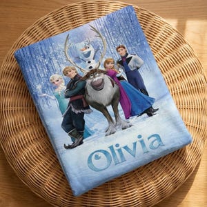 Puede incluir: Una manta azul con personajes de la pel&iacute;cula animada Frozen, incluyendo a Elsa, Anna, Kristoff, Olaf y Sven, en una escena de bosque nevado. El nombre "Olivia" est&aacute; impreso en escritura azul claro en la parte inferior.