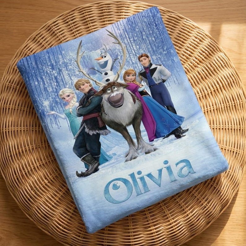 Puede incluir: Una manta azul con personajes de la pel&iacute;cula animada Frozen, incluyendo a Elsa, Anna, Kristoff, Olaf y Sven, en una escena de bosque nevado. El nombre "Olivia" est&aacute; impreso en escritura azul claro en la parte inferior.