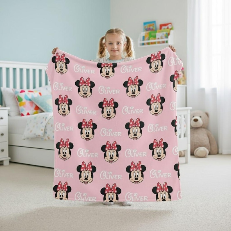 May include: A pink blanket featuring a repeating pattern of Minnie Mouse faces with red bows and the name "Oliver" in white script. The blanket is held up, showcasing its design and color.